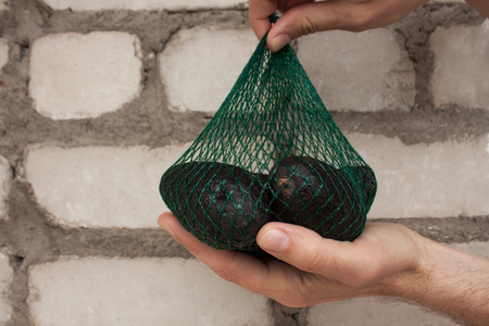 Fresh organic   avocados in a green string bag in male hands on a brick wall background, healthy food concept, copy space. Eco lifestyle, how to choose fruits and vegetables. Shopping, market.の写真素材