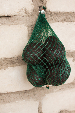Fresh organic   avocados in a green string bag on a brick wall background, creative healthy food concept, copy space. Eco lifestyle, how to choose fruits and vegetables. Shopping, market.の写真素材