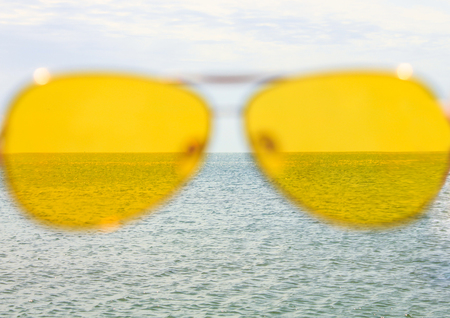 Yellow sunglasses on the sea water and blue sky background. Sunny day, happy vacation, good rest, happiness. Summer view of the sea through the glasses.の写真素材