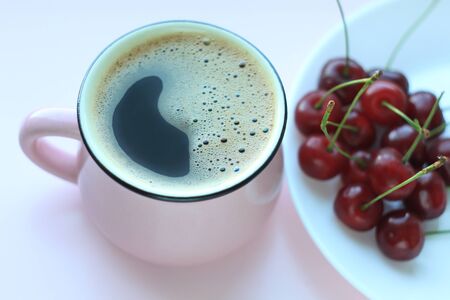 Coffee and cherries. Minimalistic concept, red cherry on white plate, fresh brewed cup of coffee on powder pink background. Appetizing breakfast, good morning, summer fruits. Top view, copy space.の写真素材