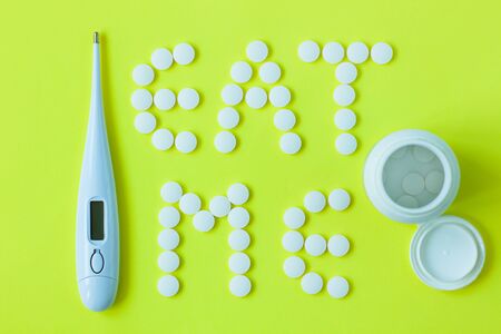 White pills in a shape of a words Eat Me with a digital thermometer and a can of drugs on a yellow background. Flat lay, top view, minimalistic style. Vitamins, treatments, dietary supplements.の写真素材