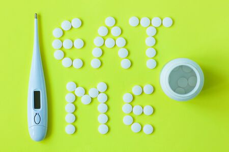 White pills in a shape of a words Eat Me with a digital thermometer and a can of drugs on a yellow background. Flat lay, top view, minimalistic style. Vitamins, treatments, dietary supplements.の写真素材
