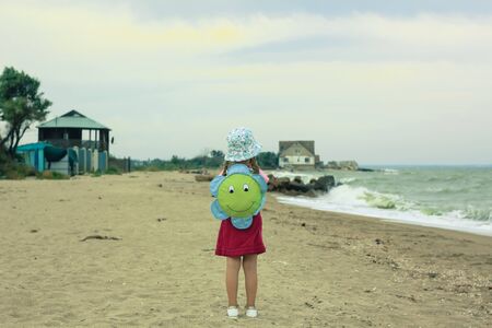 Little girl in a hat with a backpack in the form of a smiling flower on the sea sandy beach before the storm, windy cold weather, cloudy blue sky.の写真素材