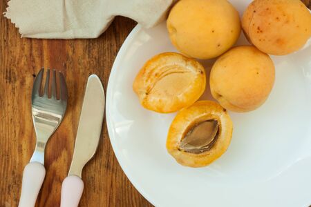 Ripe fresh apricot fruits on a white plate on the wooden background, linen towel, tablecloth, a fork and a knife. Copy space, delisious healthy food, vegan, raw. Minimalistic style.の写真素材
