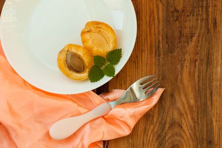 Ripe fresh apricot fruits with green mint leaves on a white plate with a fork and tablecloth on the wooden background. Copy space, delisious healthy food, vegan dishes, raw recipes.Minimalistic style.の写真素材