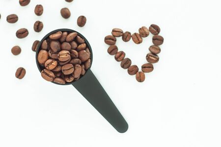 Brown roasted coffee beans in a black measuring spoon and a heart of beans on a white background isolated. Selective focus on the spoon. Coffee break, energy,freshness,great aroma.Copy space,close-up.の写真素材