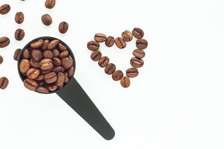 Brown roasted coffee beans in a black measuring spoon and a heart of beans on a white background isolated. Selective focus on the heart. Coffee break, energy, freshness, aroma. Copy space, close-up.の写真素材
