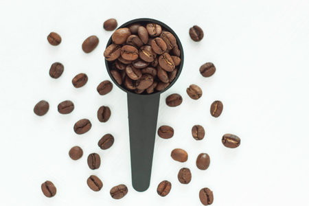 Brown roasted coffee beans in a black measuring spoon on a white background isolated. Coffee break, breakfast, energy, freshness, great aroma. Copy space, close-up.