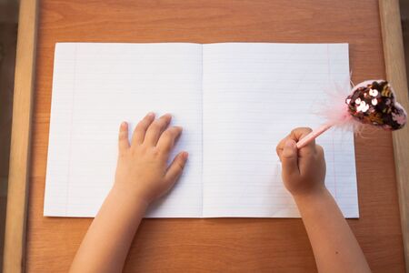 A child holds a pen in his right hand and writes in an empty notebook on a wooden desk. Back to school concept. Top view, copy space.の写真素材