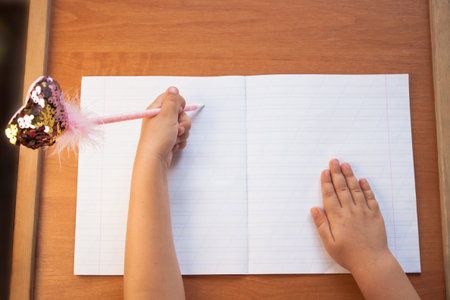 A child holds a pen in his left hand and writes in an empty notebook on a wooden desk. Back to school concept. Top view, copy space.の写真素材