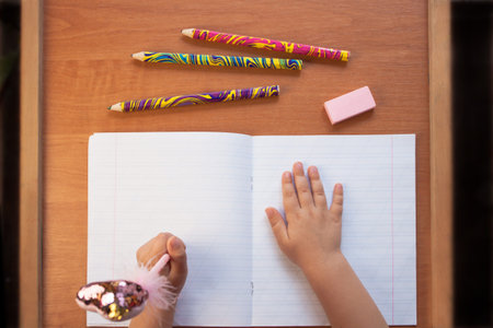 A child holds a pen in the left hand and writes in an empty notebook on a wooden desk. Stationery,...