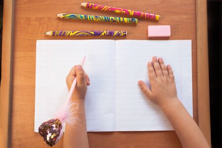 A child holds a pen in the left hand and writes in an empty notebook on a wooden desk. Stationery, colour pencils and eraser. Back to school concept. Top view, copy space.の写真素材