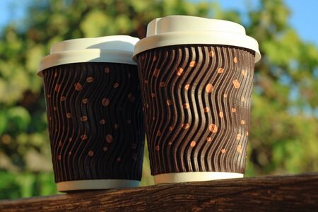 Two coffees to go on the wooden balcony. Brown corrugated cups with white lids from street shop on green nature background with bokeh lights. Lifestyle concept,take away drinks.Copy space,front viewの写真素材