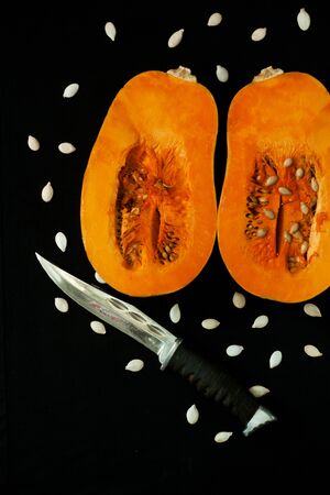 A knife and two halves of a pumpkin with seeds on a black background. Creative minimalism, dark and moody food photography. Copy space, squash top view, flat lay. Halloween concept.の写真素材