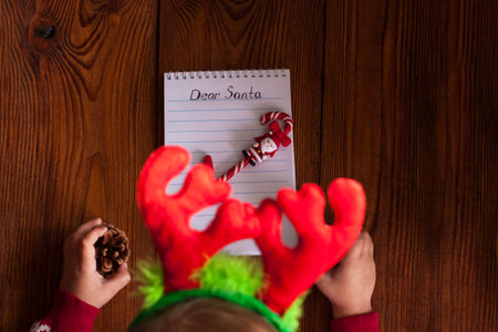 Dear Santa letter, Christmas card. Cute young girl wearing deer horns, holding a pen and writting on...