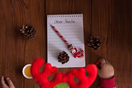 Dear Santa letter, Christmas card. Cute young girl wearing deer horns, holding a pen and writting on a white sheet on a wooden background with pine cones.Childhood dreams about gifts.New year concept.の写真素材