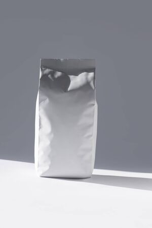 Blank foil food or drink bag on white background in minimal style with natural light shadow. Monochrome photo. Packaging with valve and seal template mockup. Metallic coffee tea retail package design.の写真素材