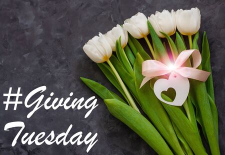 Giving Tuesday card message sign. Charity Donations concept. White tulip flowers and heart card tag with pink ribbon bow on dark background flat lay. Flower Bouquet with bokeh lights top view.の写真素材