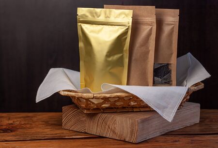 Paper and foil pouch bags with coffee beans and tea leaves in wicker basket with white textile on wooden background. Packaging for foods and goods mockup. Weight products packs with clasps and windowsの写真素材