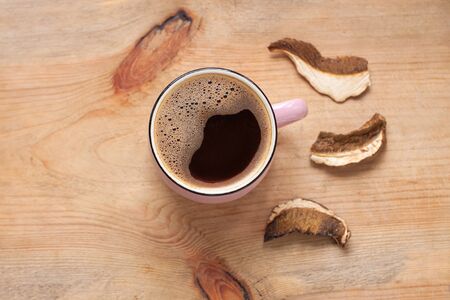 Mushroom coffee chaga superfood. Dried mushrooms and fresh brewed pink cup of hot drink with foam on wooden background top view. Healthy organic energizing adaptogen, endurance boosting food trends.の写真素材