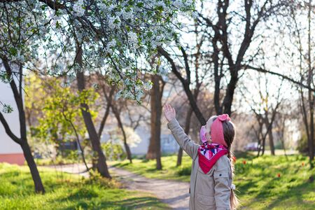 Little girl wearing pink protective face mask against COVID-19 coronavirus disease on the nature with blooming trees during quarantine.Stop epidemic.Pretty child in surgical protection on empty streetの写真素材