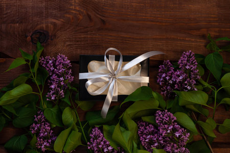 Lilac flowers, black gift box with silver ribbon on wooden background flat lay. Text place 8 March...