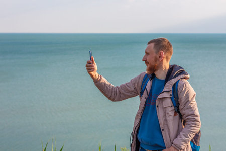 Bearded man in light raincoat on a blue sea landscape background with smartphone in hands takes seascape photography. Portrait of running young hipster guy with cell phone on nature, lifestyle photo.の写真素材