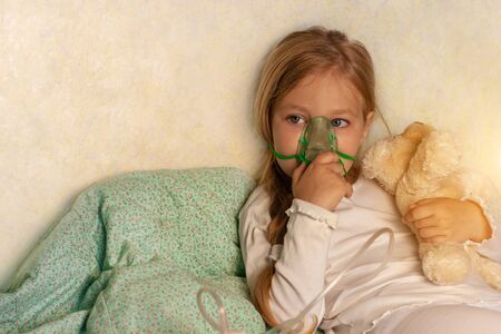 Little girl making inhalation with nebulizer on a bed at home. Sick allergic kid asthma inhaler steam cough. Child allergy concept. Respiratory diseases lungs medicine treatment.Seasonal kids rhinitisの写真素材