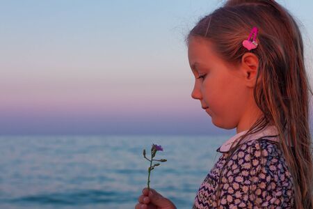 Adorable little girl child looks at the flower on beach. Portrait of a child with wet long blonde hair in dress sniffing the fragrance of wild purple flower. Amazing pink turquoise sunset on sea shoreの写真素材