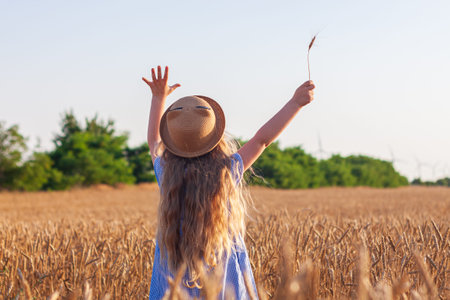 Adorable little girl in a straw hat and blue plaid summer dress in wheat field. Child with long...