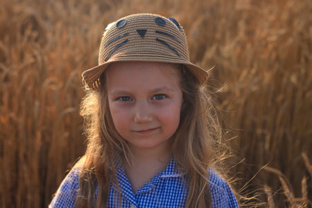 Adorable little girl in a straw hat and blue plaid summer dress in wheat field. Child with long...