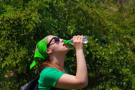 Young girl holds a bottle of water in hand and drinks. Portrait of pretty hipster in sunglasses,...