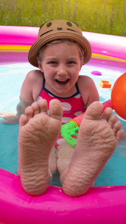 Kids feet laying on colorful rainbow inflatable swimming pool. Young girl in straw hat and pink swimsuit playing in water on a backyard. Summer active lifestyle, children swim fun leisure hot weather.の写真素材