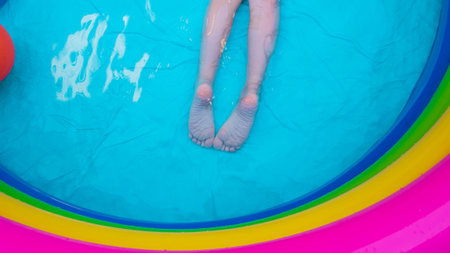 Kids feet laying on colorful rainbow inflatable swimming pool. Young girl relaxing and playing in...