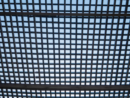 Wooden square timber lattice fence on a blue sky background. Eco wood mesh canopy with partly cloudy skies beyond. Checkered texture creative wallpaper.の写真素材
