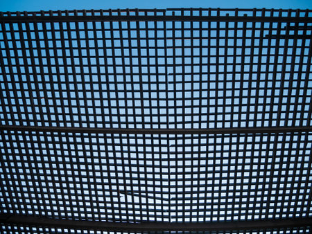 Wooden square timber lattice fence on a blue sky background. Eco wood mesh canopy with partly cloudy skies beyond. Checkered texture creative wallpaper.の写真素材