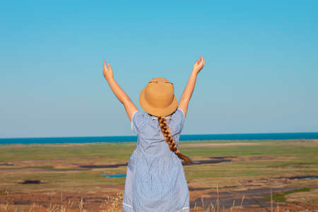 Adorable little girl in a straw hat and blue plaid summer dress in grass field with hands up to the sky. Happy child with long blonde braided hair countryside landscape. Funny walking kid outdoorの写真素材