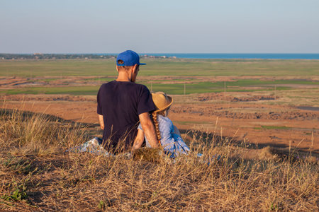 Father and daughter sitting by lakes on a nature lanscape. A little girl with long blond hair braid...
