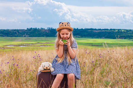 Child girl in a straw hat and dress sitting on vintage suitcase and drinking tea. Cute kid with soft dog toy looking at the cup in hands on nature lanscape background. Adventure concept in retro styleの写真素材