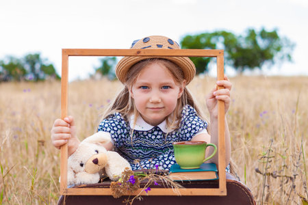 Child girl in a straw hat and dress sitting behind vintage suitcase with cup tea. Cute kid with soft dog toy looking in wooden frame in hands on nature lanscape background.Creative concept retro styleの写真素材