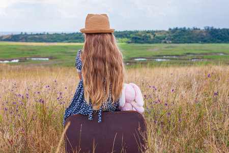 Child girl with long hair in straw hat and dress sitting on vintage suitcase and taking a picture. Cute kid with soft toy looking at nature lanscape background. Adventure travel concept in retro styleの写真素材