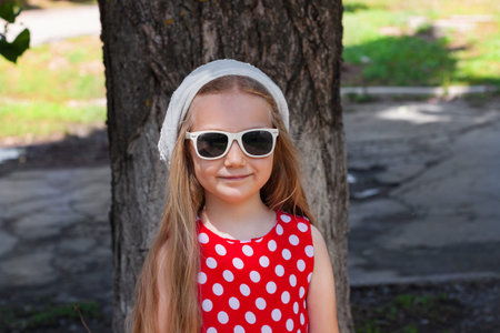 Beautiful happy girl in a red polka dot dress and white beret smiling on green street background. Cute joyful child with long blonde hair in sunglasses and hat on natural backdrop outdoor.の写真素材