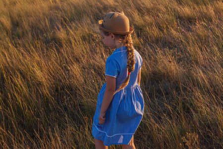 Adorable little girl in a straw hat and blue plaid summer dress in grass field with wild flowers in hands. Happy child with long blonde braided hair countryside landscape. Funny walking kid outdoorの写真素材