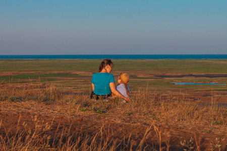 Mother and daughter sitting by lakes on a nature lanscape. A little girl with long blond hair braid hugging with mom outdoor. Loving child embrace her mommy. Mother's Day family greeting card facelessの写真素材