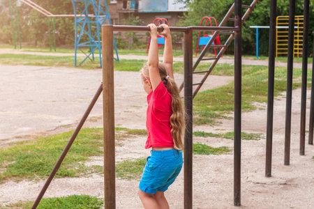 Girl hanging on horizontal bar. Child doing motion exercises and playing on playground outdoor....