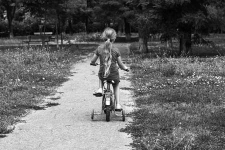 Child riding on bicycle. Blonde smiling girl on bike on asphalt road in summer. Active lifestyle leisure, physical training on sports stadium. Happy kid bicyclist. Black and white street photo.の写真素材