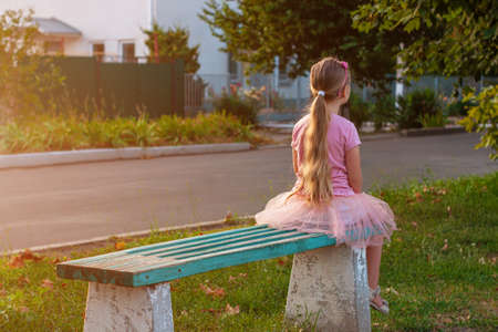 Cheerful little girl with long blonde hair in pink tulle skirt sitting alone on a bench on empty street on sunset. Happy dreamy cute child on nature background. Kids dress fashion. Greeting card.の写真素材