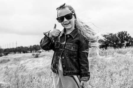 Adorable little girl in denim jacket, sunglasses, blue plaid dress in grass field. Happy stylish long blonde hair child on countryside landscape. Cute kid walking outdoor rural road trip. Monochromeの写真素材