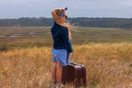 Little blonde girl in denim jacket, black sunglasses, blue dress with vintage suitcase in wild grass field. Stylish hitchhiker child with long hair countryside landscape trip. Kid walking outdoor roadの写真素材