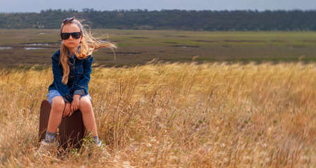 Little blonde girl in denim jacket, black sunglasses, blue dress with vintage suitcase in wild grass field. Hitchhiker child with long hair countryside landscape trip. Kid walking outdoor road bannerの写真素材